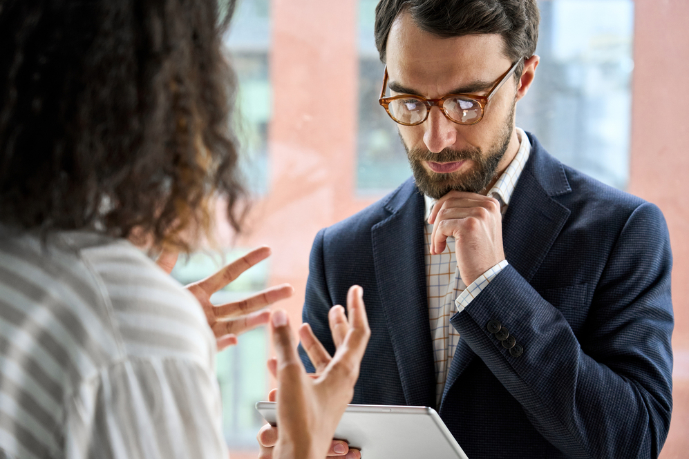 Serious executive middle aged ceo bearded boss businessman using looking at tablet device listening to female manager negotiating explaining discussing project report at office. Over shoulder view.